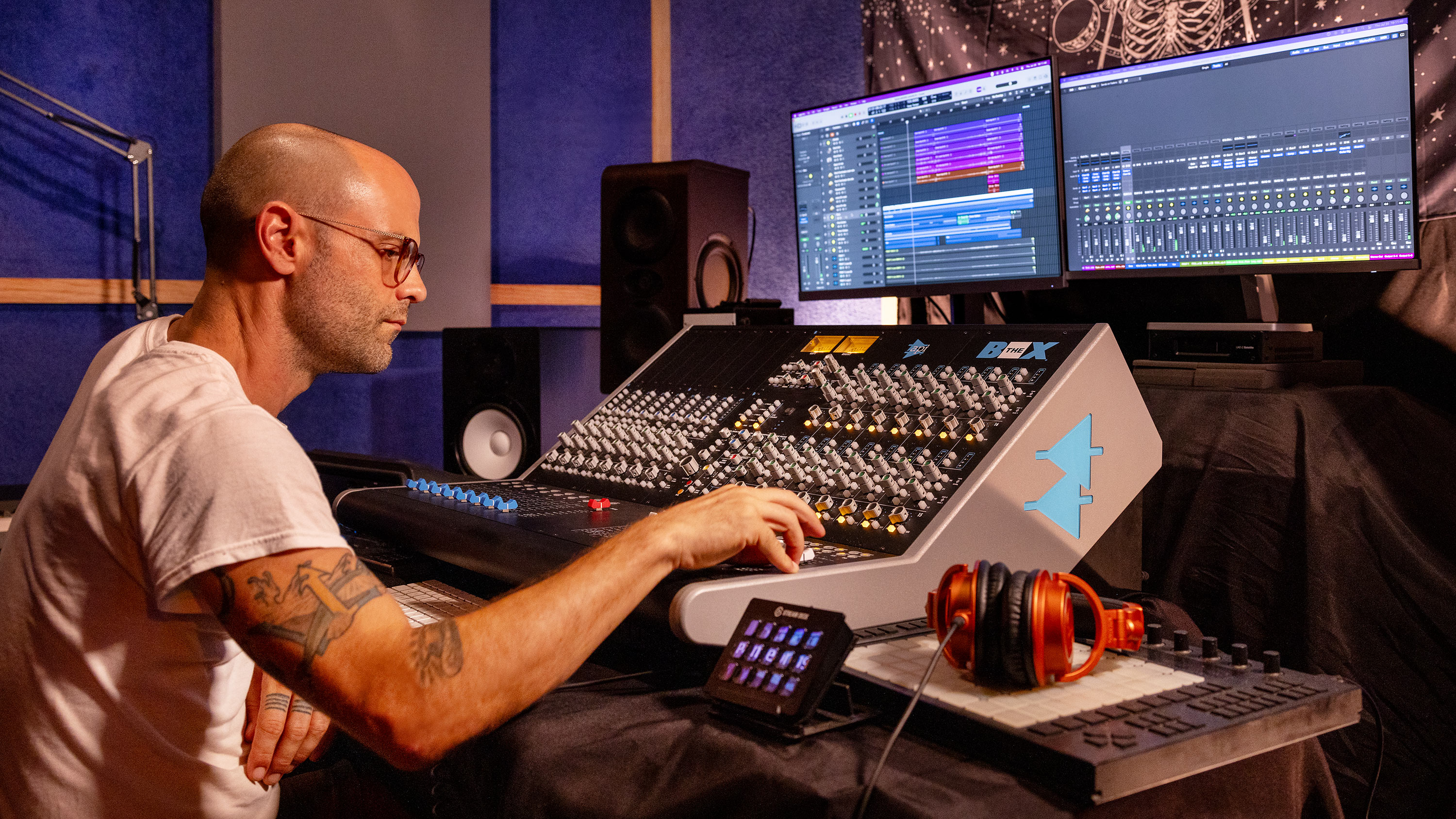 Producer working at professional mixing console with dual monitors showing DAW